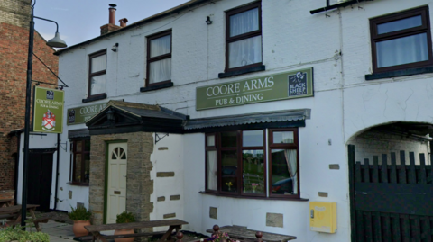 A traditional white‑painted pub called the Coore Arms, with dark brown window frames, a small stone porch at the entrance, and hanging signs for “Coore Arms Pub & Dining.” Wooden benches and potted plants sit outside on a quiet village street.