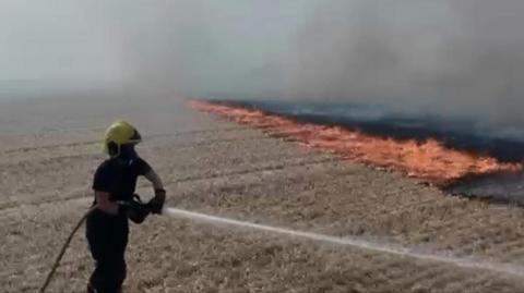 Someone in dark fire officer uniform, with a yellow helmet and a face mask, points a fire hose at a dry brown field, with water blasting out. To the right of the frame are red flames flicking on the field.