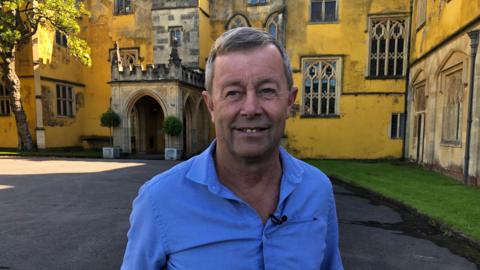 Norman Routledge smiles at the camera in front of the yellow painted entrance to the 18th century mansion at Ashton Court