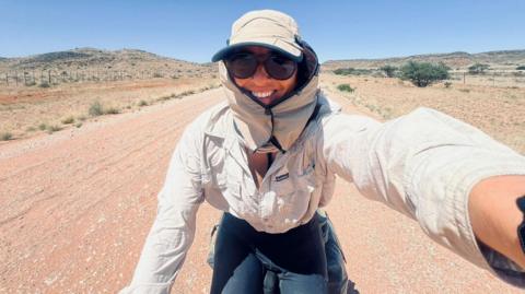 Ellie Mitchell-Heggs on her bike. Her head is covered with a hat with side panels and she is wearing a sand coloured shirt and black leggings. She is cycling on a sandy dirt road with dry greenery each side. There are hills in the background and she is smiling as she takes the selfie.