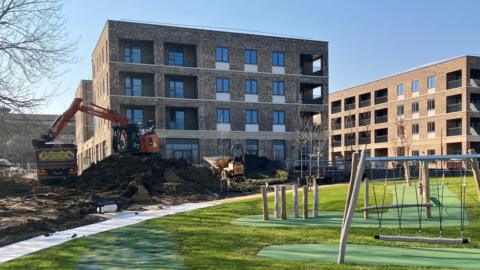 A grassy play area with swings sits in front of modern apartment buildings, while construction work with an excavator is underway on the left side of the scene.