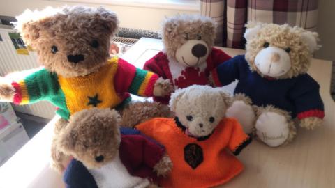 Five teddy bears of varying sizes sit together on a desk. They all wear different knitted shirts in different colours.