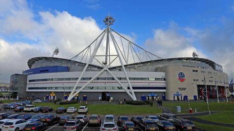 A football stadium viewed from a car park with a large white sail-like structure over a curved stand. The Bolton Wanderers club crest is visible on the side of the building