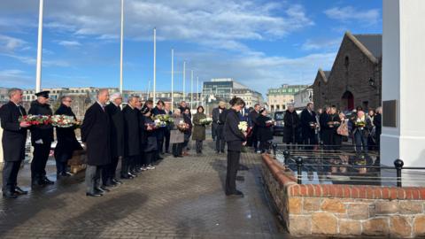 People standing around a memorial in all black holding flowers with their heads down in respect.