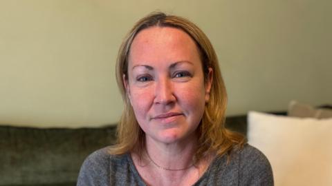 A woman with shoulder length blonde hair is looking into the camera and smiling slightly. She is wearing a dark grey round neck top with a fine gold chain necklace. the background is out of focus, but she is sitting on a dark coloured sofa with cream cushions. 