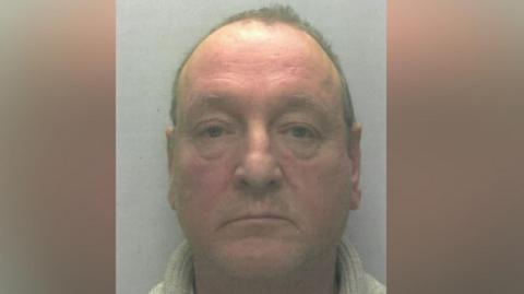 A police mugshot of a man in his sixties, with a reddish face and short grey hair. He is wearing a grey turtleneck jumper.