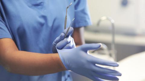 A dentist puts on blue gloves while holding the metal tools needed for an examination