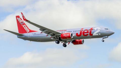 A red and white Jet2 aeroplane is flying through a blue sky with large white clouds. The aircraft’s landing gear is down and the airline name “Jet2.com” is clearly written on the side and tail.