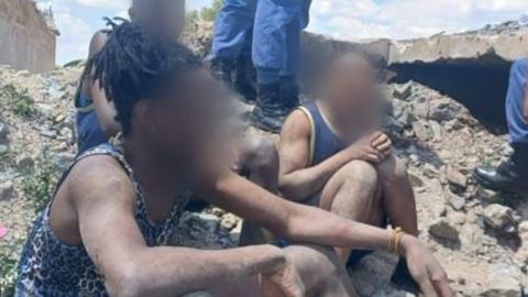 Two men with their faces blurred out sit on a rocky ground. They are covered in dust and are wearing vests.