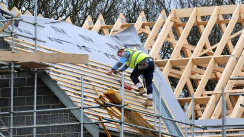 A roofer is wearing a high-res jacket and he is carrying out building work on the roof of a property. 