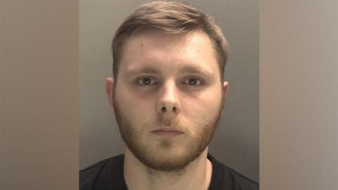 Joel Dever is pictured in a police mugshot. He has short light brown hair and a beard and is wearing a black top.