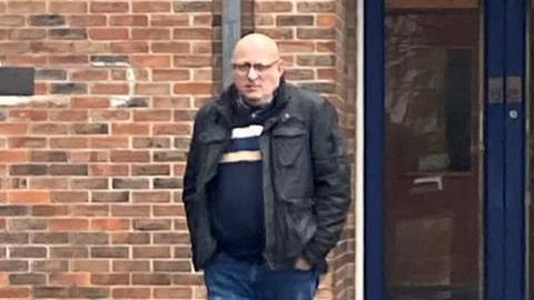 Jonnathan Hicks outside court. He has a bald head and glasses, and is wearing a blue jumper, jeans and a black open jacket.