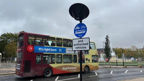 A burgundy, yellow, gold and red painted bus travelling in a bus lane. There is a post on the path with a blue circular sign with a motorcycle, cycle and bus image on. It also reads: "taxi", in white. Below it is another sign, which is white and has black writing on reading: "and authorised vehicles". There is a fountain in the background. The sky is overcast. 