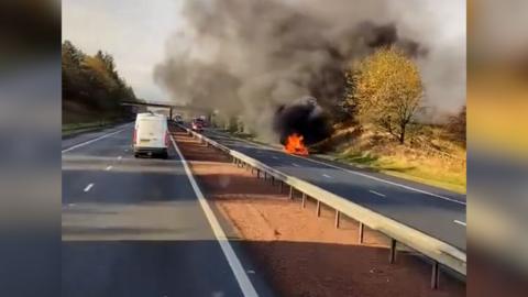Dashcam footage of a car in flames at the side of a motorway with black and grey smoke.