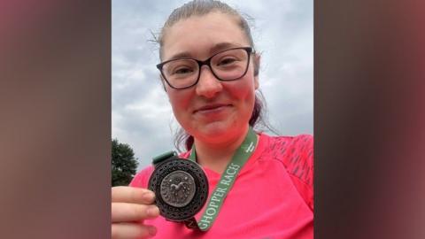 Asher Cooper who is taking a selfie and looking directly at the camera and smiling. She has a medal round her neck and is holding this up and showing the camera. She is wearing a hot pink running top and is wearing glasses.