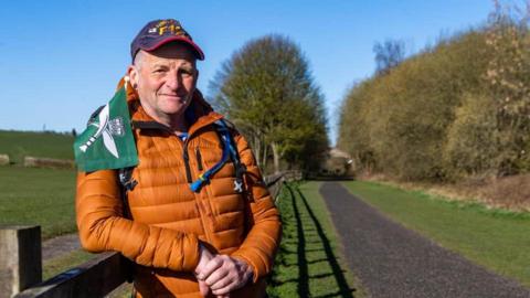 A man in an orange puffer jacket and blue and red hat stands beside a country path. A green Gurkha flag rests on his right shoulder. Trees and hedges stand behind him.