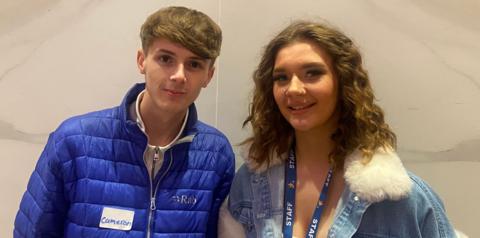 A young man wearing a blue coat, with short blonde hair, and a young woman with wavy brown hair and a denim jacket are standing in front of a wall