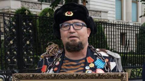A man wearing an old-style Russian hat with a Soviet badge holds the top of a framed picture and wears a jacket crammed with medals