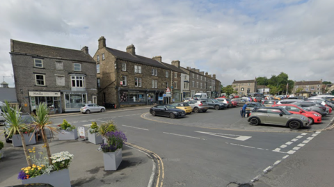 A town square with a parking area filled with various cars, surrounded by stone buildings with shops on the ground floor. The foreground features decorative planters with flowers and small palm plants along the edge of the road.