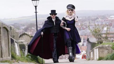 A man and a woman dressed in gothic clothing walk arm in arm through the graveyard near Whitby Abbey