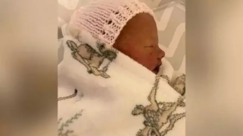 A newborn baby in sleeping in a cot wearing a white beanie and wrapped in a white blanket. 