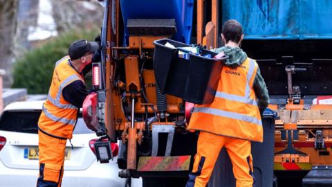 Two men in orange high-vis clothing empty rubbish from bins into a lorry in a residential street