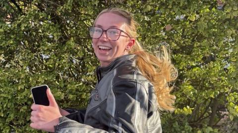 A woman wearing grey and black bike leathers is holding a mobile phone and running. She is wearing glasses and has blonde hair.