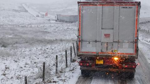 A lorry which is being driven on the B6276. The road is covered in snow and there are many vehicles which look like they have been abandoned.