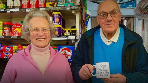 Pauline and Derek Hughes standing inside their shop in Cotteridge. Derig, wearing glasses, a blue jumper and dark blue jacket is holding a mug, while Pauline, on the left is wearing glasses and a pink jacket