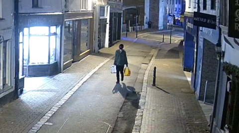 This is an image of a person with a black face covering. They are holding a large silver ladder and a yellow bag. They are walking on a stretch of street. It is dark.