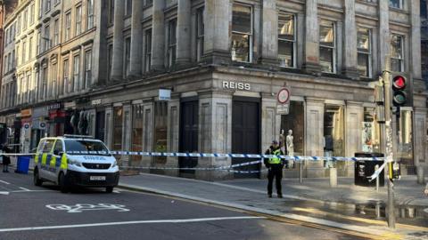 Police cordon blocks a city street beside a large stone building with columns, as officers stand guard and vehicles are stopped nearby.