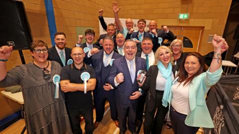 People in suits and pale blue rosettes with hands in their air celebrate