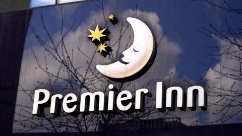 A photo of a large Premier Inn sign which has the company's moon logo above its name. The sign is a reflective surface and reflections of trees can be seen against a cloudy but blue sky.