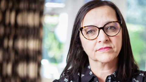 Figen Murray with long dark brown hair wearing tortoise shell glasses and a black shirt with white patterns on it. She has her head tilted slightly and is looking impassively at the camera. A curtain is pulled back and a window showing a garden in the blurred background. 