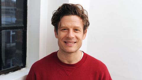 James Norton looks at the camera, smiling. He is wearing a red jumper and standing in front of a white wall next to a black framed window.