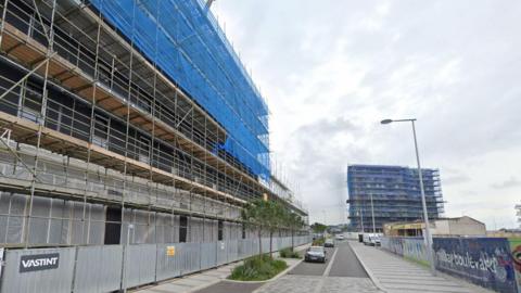 A road in Plymouth with cars driving down it and blocks of housing being built along the roadside with lots of scaffolding set up.