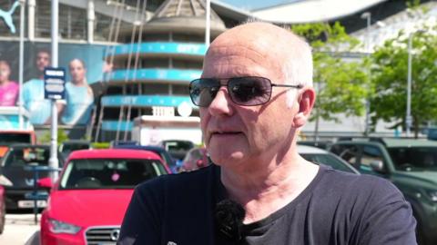 A man with shaved white hair, looking slight off camera, he is in the car park of Manchester City stadium as the ground can be seen behind him. He has a black T-shirt and has sunglasses on. It is a head and shoulders shot.