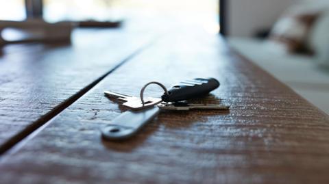 A set of house keys on a key ring with fobs laid out on a wooden table. Sunlight pours in from a window.