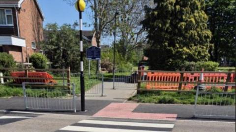 A pedestrian crossing leading to a park with a blue sign at the entrance