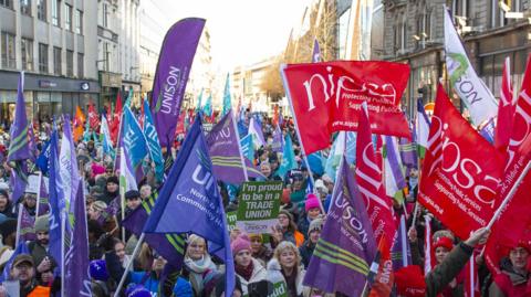 Hundreds of people from different trade unions stand in a Belfast street waving red, blue, purple and green flags