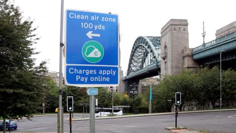 A blue sign reading Clean Air Zone 100 yards, charges apply, pay online points to the left. Newcastle's Tyne Bridge is in the background. It is a green metal arch structure.