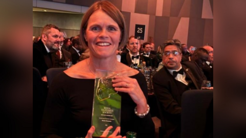 A woman in a black off-the-shoulder dress holding up an oblong glass award. It is engraved with Royal Television Society, then in smaller letters says Midlands Awards 2024, Breakthrough on Screen Unscripted, Nicola Goodwin, ˿. The woman has short brown hair in a bob. Other guests in black tie can be seen at tables behind her in an events venue.