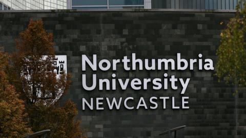 The outside of a Northumbria University building which is a large, grey brick structure. Northumbria University Newcastle is written on the wall in large silver letters with the crest on the left, which is obscured by a tree with orange leaves. 
