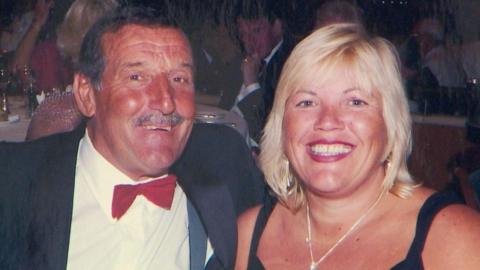 Colin, with dark hair and a moustache, is smiling at the camera, wearing a red bow tie, white shirt and black blazer. He is sat next to his partner Melanie, who has blonde shoulder-length hair with a fringe and is smiling with a plum coloured lipstick