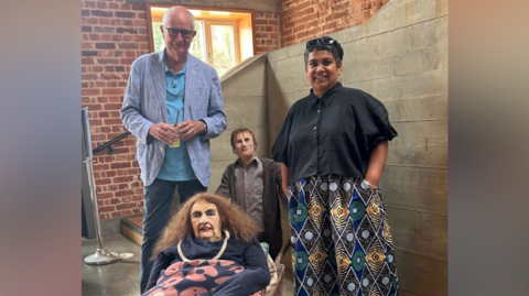 Jim Broadbent and Devi Singh stand together with two of his sculptures placed in between them. Jim has a bald head and wears a blue jacket with a blue polo top underneath and navy jeans. Devi wears a black top with patterned wide leg trousers. One of the sculptures sits in a wheelbarrow and has long brown hair with a navy jumper and blue shirt with pink flowers on it. The other sculpture holds the wheel barrow behind and has short hair, wears a shirt and a cardigan. 