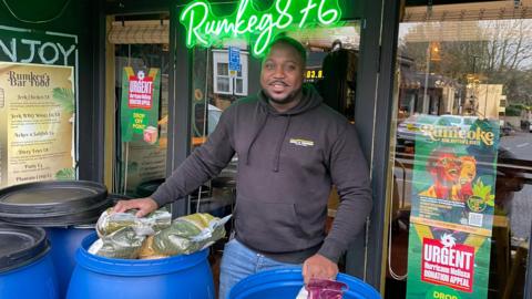 A man stood with his hands on two blue barrels which are filled with big bags of dry food. He has short dark hair and a beard and is wearing a black hoodie and blue jeans. 