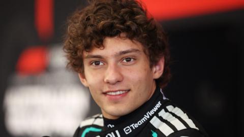 A young motorsport driver with curly brown hair, dressed in a sleek black racing suit accented with white and teal details. Sponsor logos are visible across the suit, including one on the collar. 