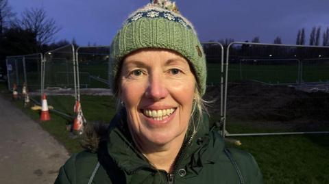 Close-up of Julie Norman who is smiling directly at the camera. She is wearing a green coat and woolly hat and is standing in front of metal barriers in a park. It is early morning and the sky is a dark purple.