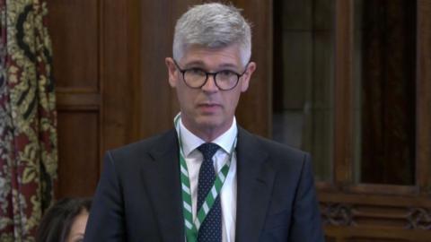 Al Pinkerton, the Surrey Heath MP, talking in a debate at Westminster Hall in Parliament. He is wearing a suit and glasses and has a green and white ribbon around his neck.