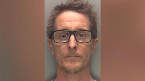 A police mugshot of a man with short brown hair and thick black-rimmed glasses and a short moustache and beard, looking into the camera.